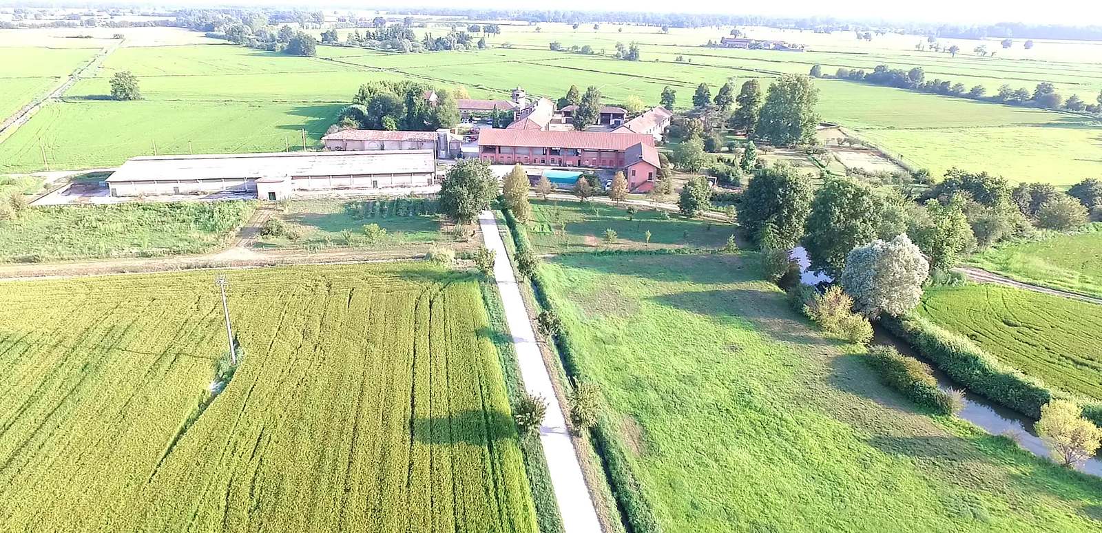 La cascina vista dal drone in fondo alla strada bianca, tra un campo di riso e una roggia